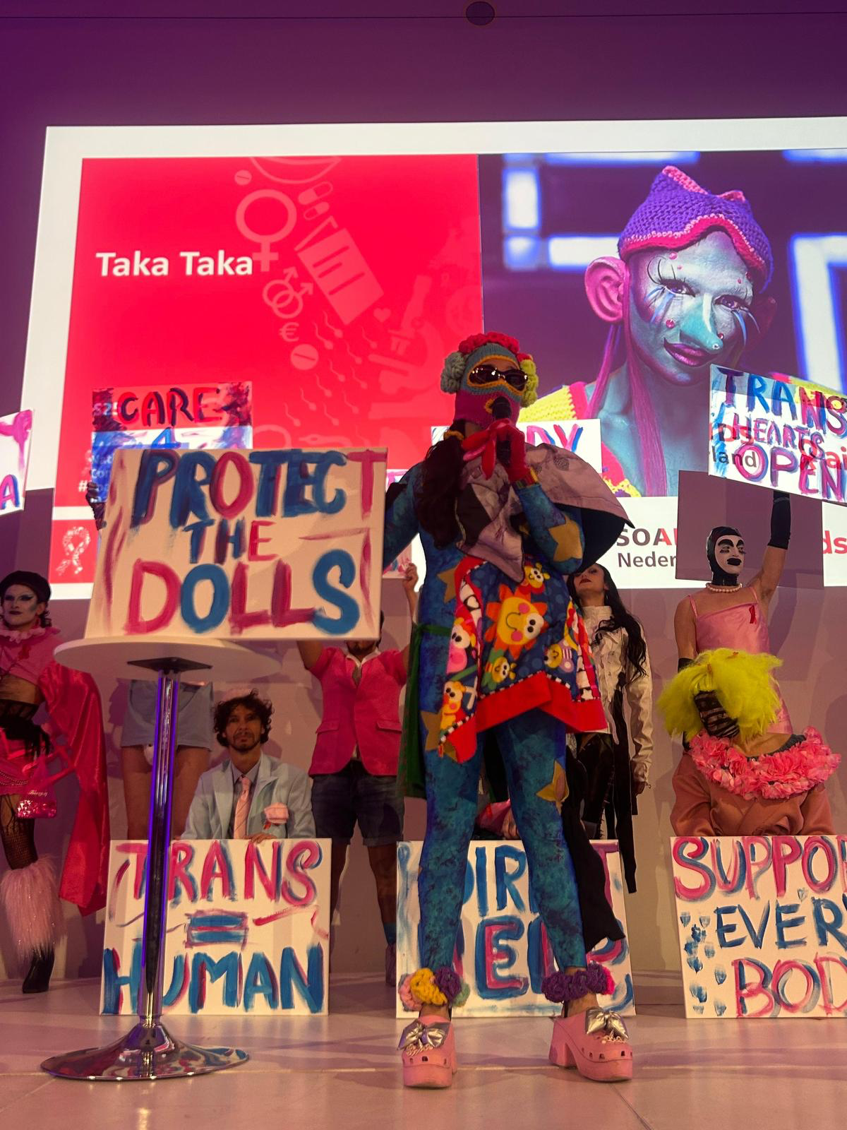 Image description: Taka stands at the front wearing a colourful costume. Behind them are members of the House of Hopelezz and the House of Løstbois, holding protest signs with handwritten messages focused on trans awareness. The group is dressed in drag, including a mix of trans men and trans women. The photograph was taken at the main national STDs conference in the Netherlands, held in The Hague on 1 December, World AIDS Day.