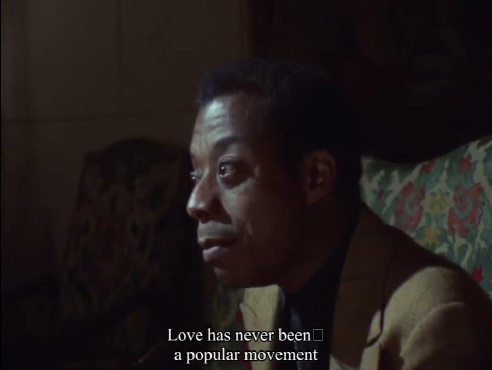 Image description: A colour film still from Meeting the Man: James Baldwin in Paris (1970), sourced by Emma Ajdari & gervaise alexis savvias for love and innocence in the thrall of carcerality [The image shows James Baldwin looking past the camera with a reflective expression. Subtitles on the image read: “Love has never been a popular movement.”]